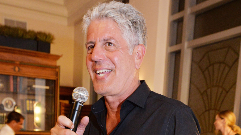 Anthony Bourdain speaking into a microphone inside a restaurant