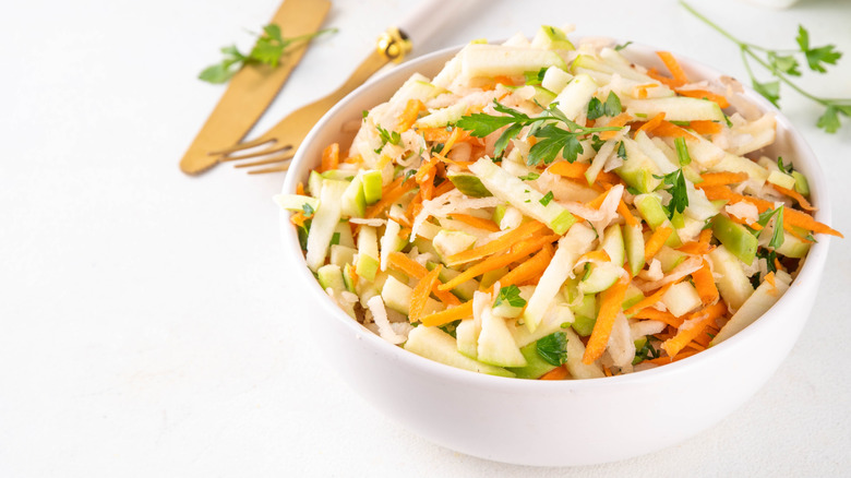 A bowl of apple coleslaw on a white surface with wood utensils nearby.
