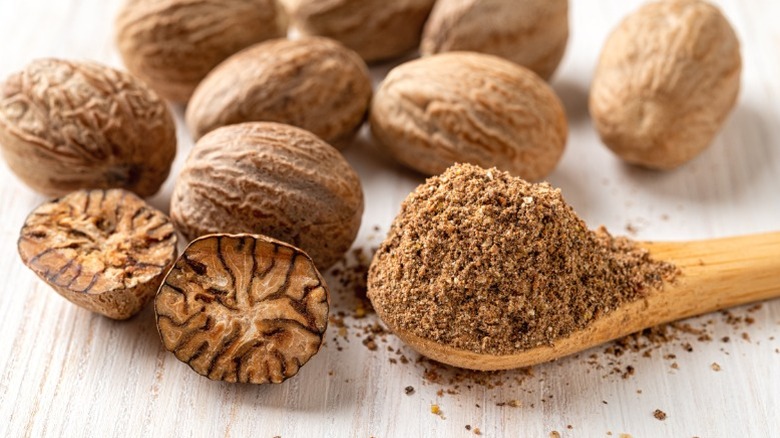 Powdered nutmeg on a wooden spoon surrounded by whole nutmeg.