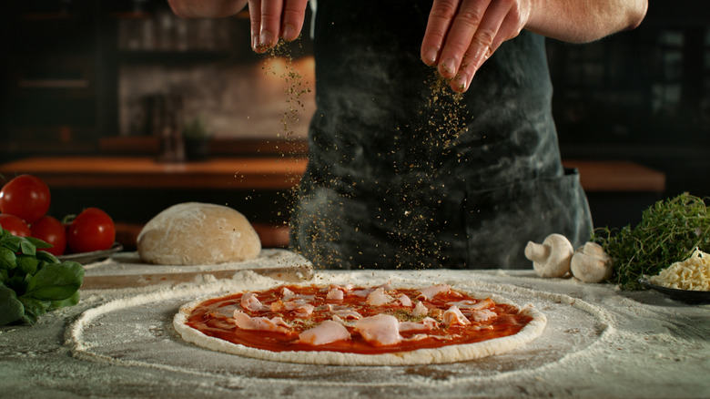 A chef sprinkles seasonings onto an uncooked pizza with sauce