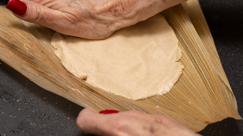 Hands pressing masa dough into a corn husk to make tamales