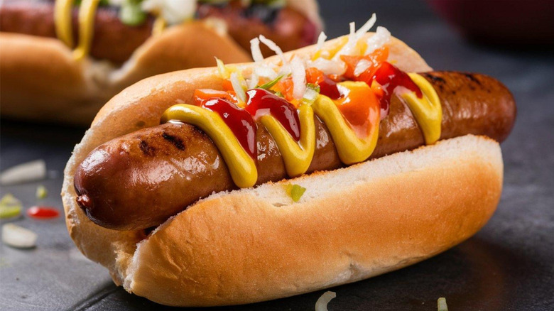 Grilled hot dog on a bun with mustard, ketchup, and onions on a black surface