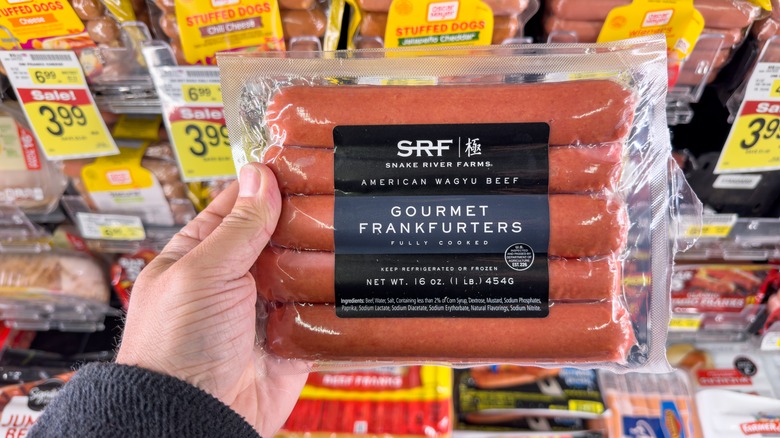 Hand holding a pack of five wagyu beef hot dogs in front of a store selection of other hot dogs