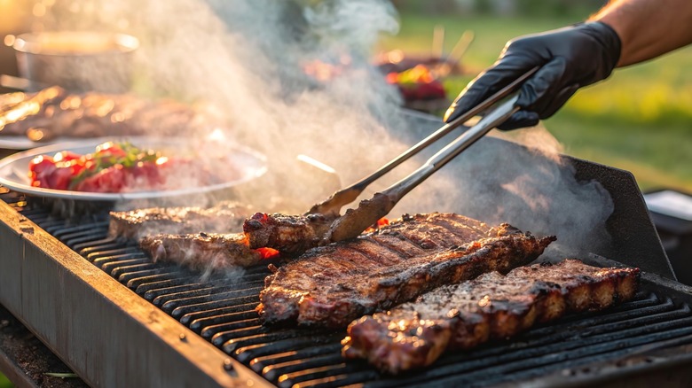 A person's gloved hand uses tongs to position meat on a grill