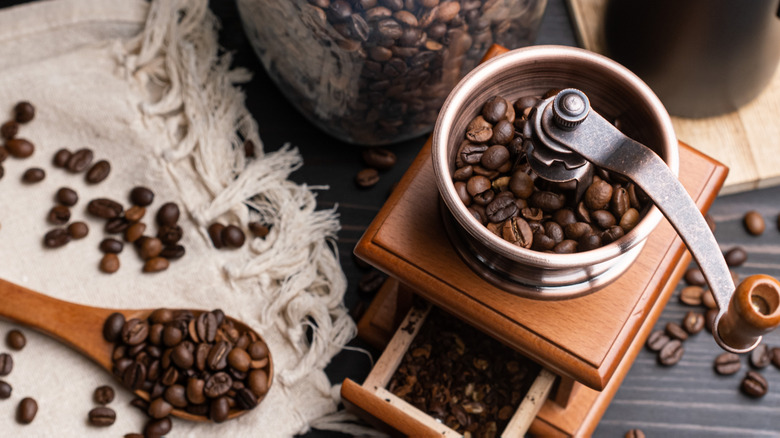 Overhead view of coffee beans in a grinder near loose beans, a wooden spoon, and placemat or dish towel