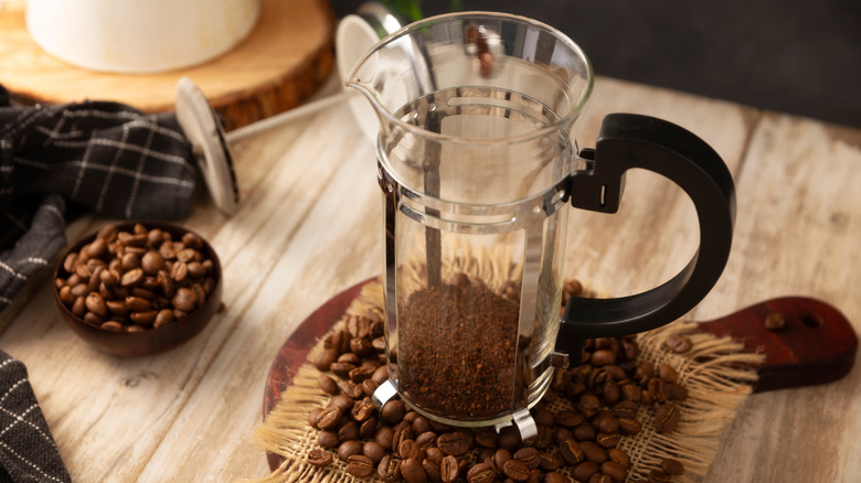 A French press carafe with ground coffee sits on a pile of coffee beans on a wooden surface.
