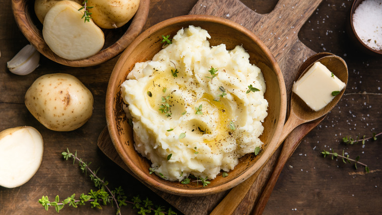 A wood bowl of mashed potatoes near sliced potatoes, butter, and salt.