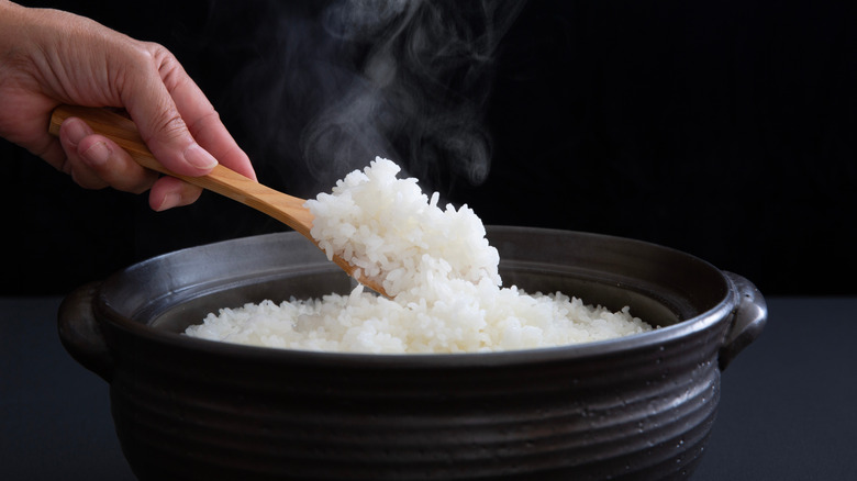 A person scoops cooked white rice from a black pot with a wooden spoon