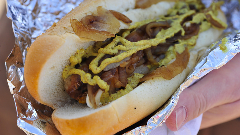 Hand holding hot dog with cooked onions and mustard wrapped in foil