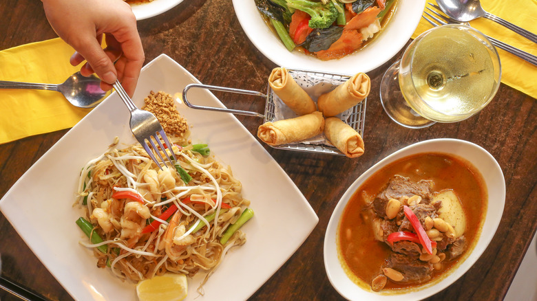 The image shows a hand with a fork reaching for the plate of Pad Thai prawns, a metal basket with 4 spring rolls, a red curry, a bowl of stir fried vegetables, and a glass of white wine.