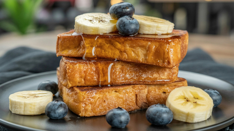 Three thick pieces of French toast with bananas, blueberries, and syrup on them