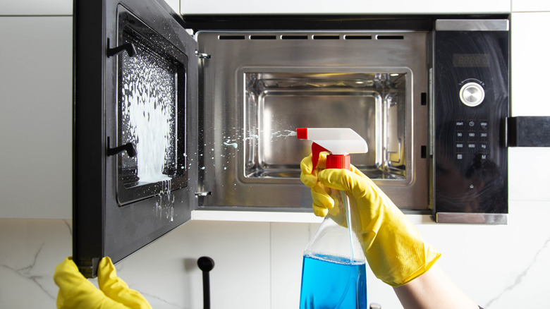 Someone wearing rubber gloves and spraying a microwave door with cleaner