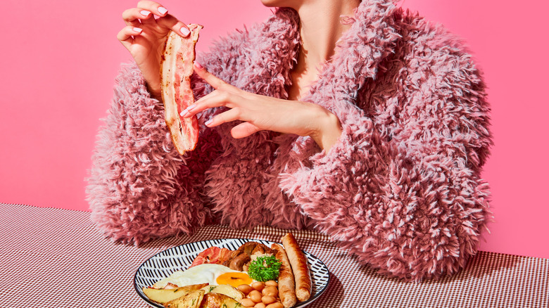 A woman in a pink furry coat holding bacon and seated by plate of breakfast foods
