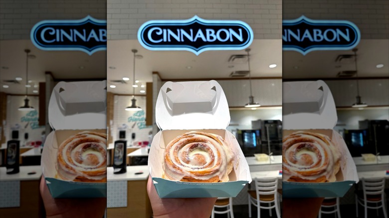 Big cinnamon roll in cardboard box held in front of sign in Cinnabon store