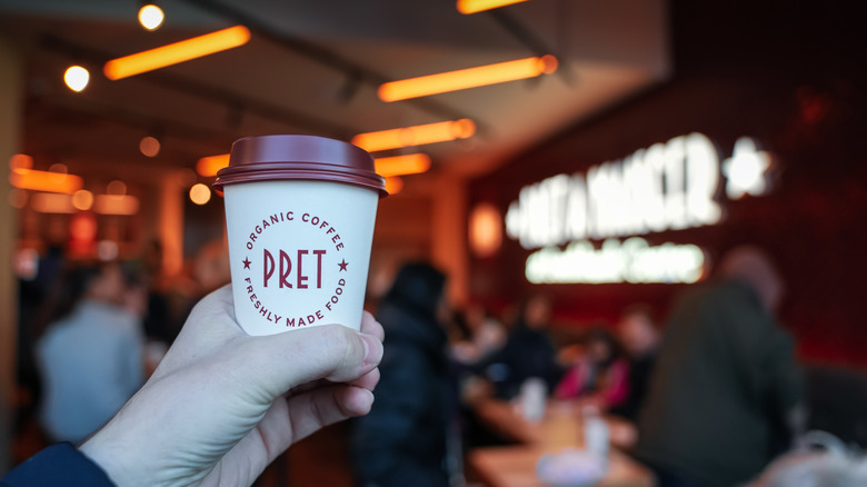 Hand holding takeout coffee paper cup with Pret a Manger logo and bakery in background