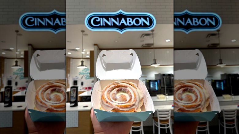 Big cinnamon roll in cardboard box held in front of sign in Cinnabon store
