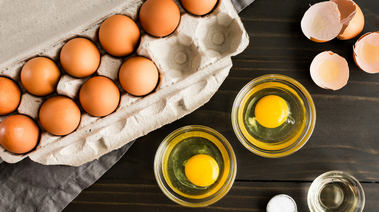 Carton of brown eggs with two missing, with two small bowls with one cracked egg in each and the shells on the side on a dark wood surface