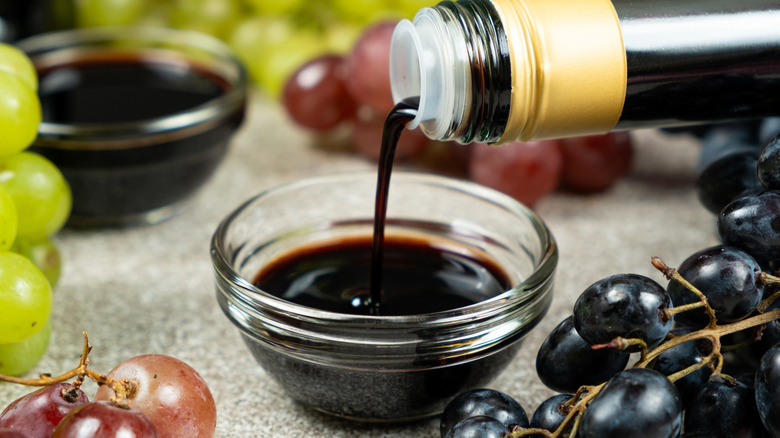 Dark balsamic vinegar being poured from a bottle into a small glass bowl surrounded by red, green, and black grapes