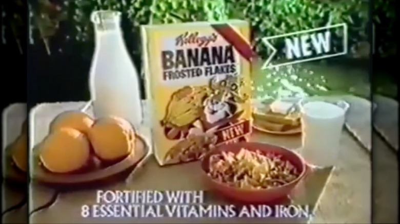 Box of Banana Frosted Flakes sitting next to bowl of cereal, bottle of milk, and whole oranges from a commercial