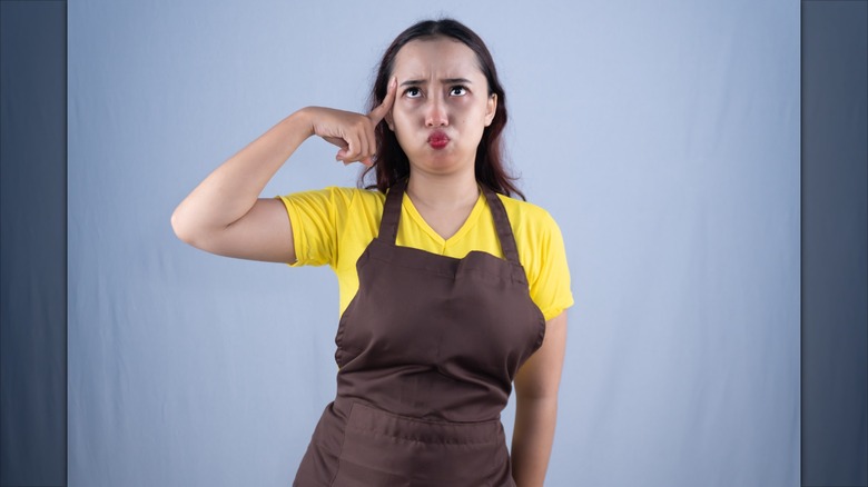A woman wearing a yellow shirt and brown apron pointing at her temple while making a confused expression