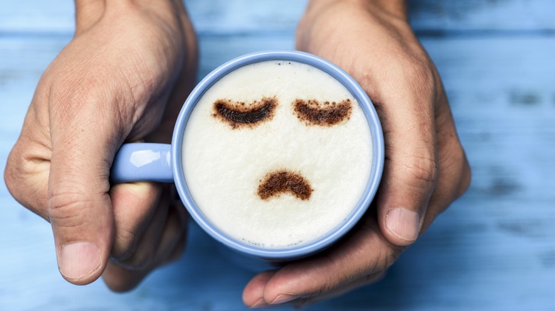 Hands holding a blue cup of cappuccino with a sad face drawn with cocoa powder on the milk foam