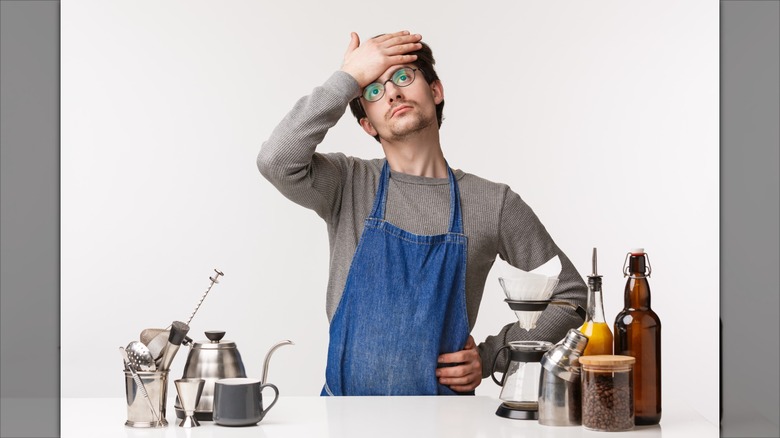 Man in an apron with his hand over his forehead, surrounded by coffee making equipment.