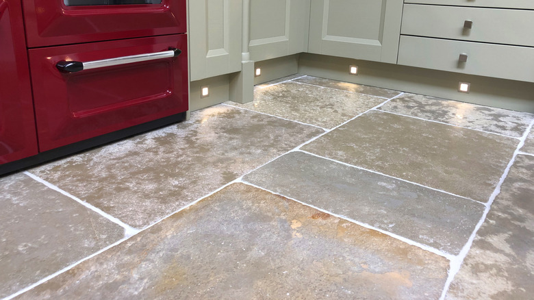 A closeup of a stone kitchen floor with gray shelves and red stove in the background