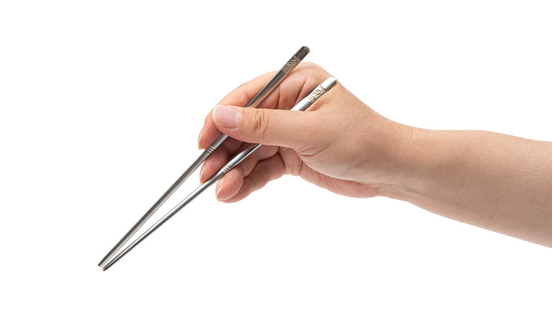 A hand holding two metal chopsticks in front of a white background
