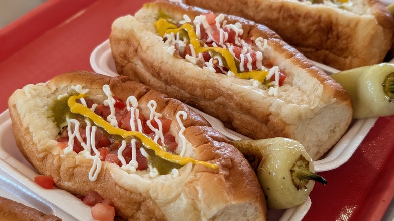 Sonoran hot dogs garnished with mustard, mayo, green salsa, and roasted peppers in to-go containers