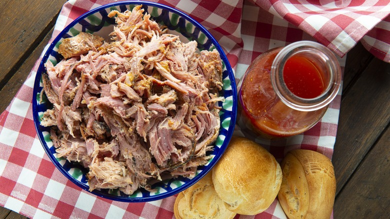 Pulled pork in a decorative blue bowl with a side of vinegar barbecue sauce and rolls on a red checkered napkin