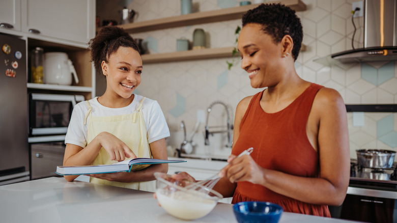 A mother whisking batter while her daughter reads from a cookbook.