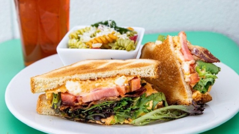The Hungry Peach Southern BLT on a white plate with a bowl of pasta salad and an iced tea in the background