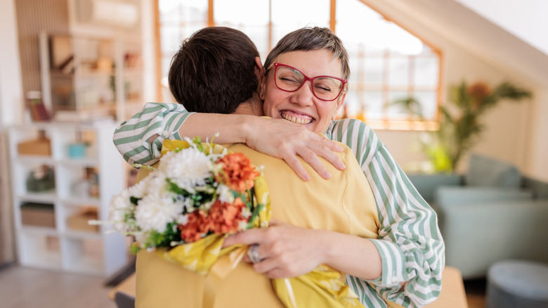 Joyful senior mother embracing her son tightly while holding a colorful bouquet of flowers in a bright, cozy living room. The genuine smiles and warm atmosphere express love, gratitude, and deep family connection.