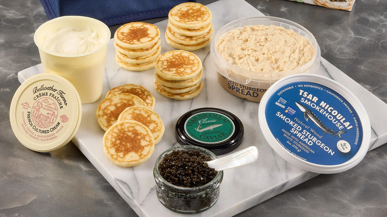 A marble cutting board with stacks of round blinis, a jar of caviar, and containers of white spreads