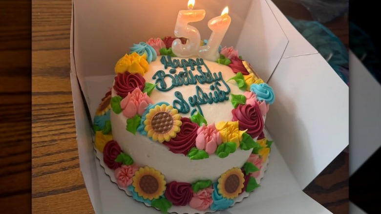 White-frosted round birthday cake with piped flowers and sunflowers with a lit 51 candle on top