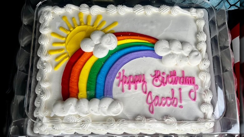 White-frosted sheet cake with a sun and rainbow design and pink lettering that reads "happy birthday jacob"