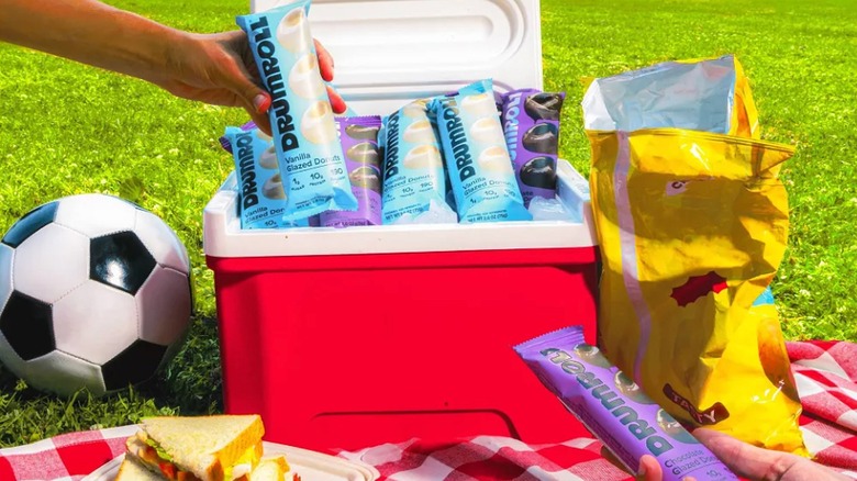 People reaching for a package of Drumroll Mini Donuts from a red cooler sitting on the grass next to a soccer ball