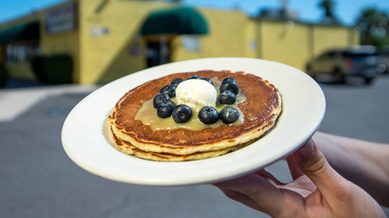 Hand holding up a plate of pancakes with blueberries and butter outside in front of Joe's Diner