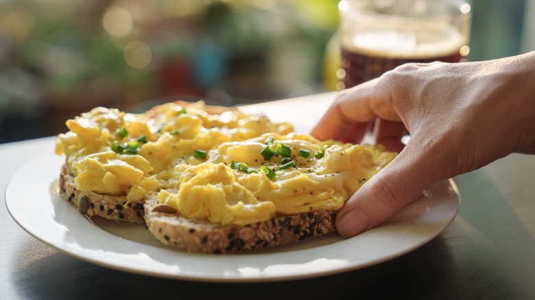 A cropped image of a hand holding a piece of toasted bread with scrambled eggs on top