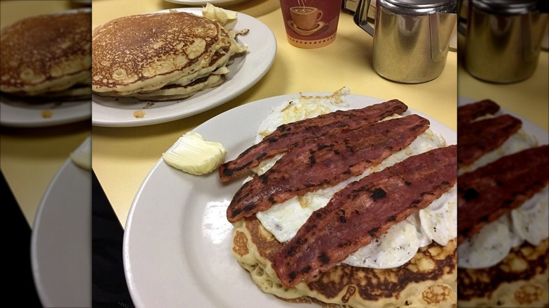 Plate of pancakes topped with egg and bacon, with another pancakes plate in background at Johny's Luncheonette