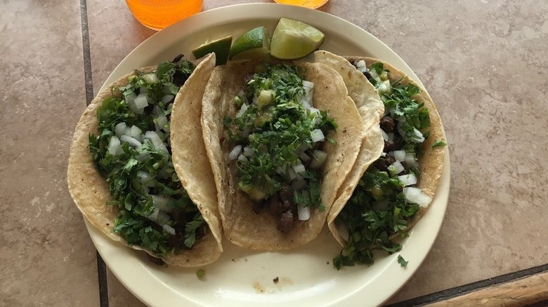 Three carne asada tacos in corn tortillas on a white plate on a taupe tile surface