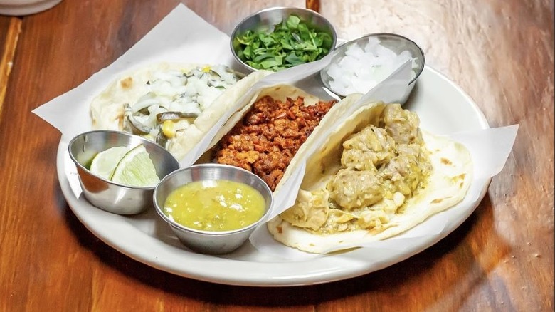 Three soft tacos and four stainless-steel ramekins of condiments on a white plate