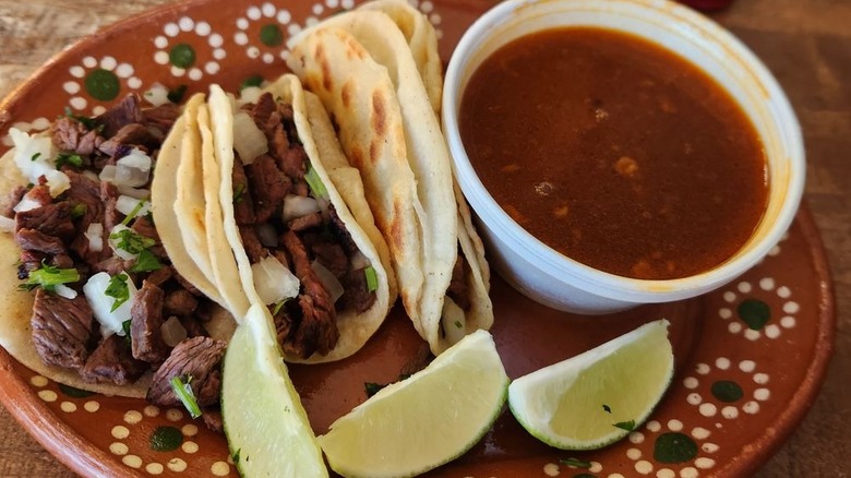 Three steak tacos with cliantro and onion on the left side of a brown plate decorated with a dot pattern with a plastic ramekin of consumme on the left side and three lime wedges on the forefront of the plate