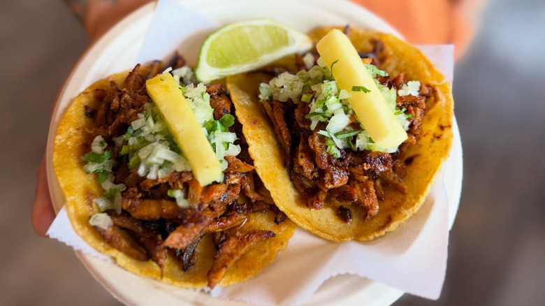 Someone holding a white paper plate with two al pastor tacos on flour tortillas