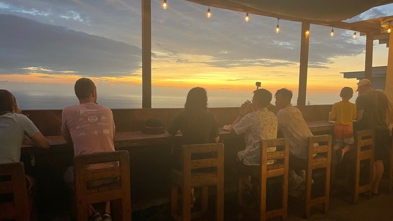 Six wood barstools lined up on a counter on a wooden deck that overlooks the Pacific Ocean at dusk