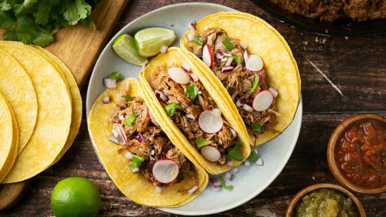 Three tacos on double corn tortillas filled with shredded meat and topped with cilantro, chopped onions, and sliced radishes and garnished with two lime wedges on a gray plate set on a dark wood surface; on the left, a cutting board with cilantro, corn tortillas, and a whole lime; on bottom right corner, a brown porcelain ramekin of red salsa and another ramekin of green salsa