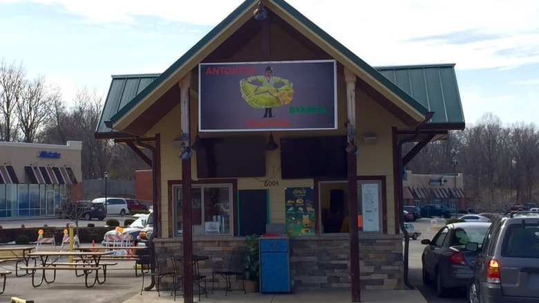 Small walk-up or drive-thru food stand with green metal roof, beige siding and brown and brick trim in a shopping plaza parking lot; a few picnic tables are set up on its left and there are two vehicles waiting in the drive-thru area