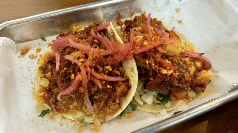 Two al pastor tacos on flour tortillas on a rectangle-shaped metal tray lined with wax paper; topped with pickled onions, pineapple, red onion, cilantro, and pork rind crumbs