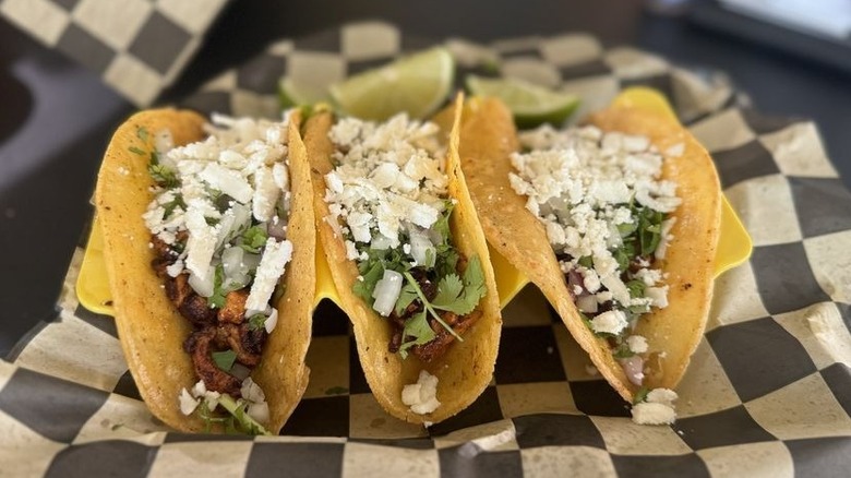 Three tacos on corn tortillas set on a yellow taco stand on top of a black-and-white checkerboard-lined tray; each is filled with meat and topped with onions, cilantro, and cotija cheese