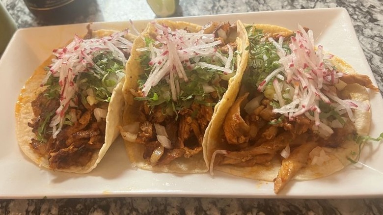 Three el pastor tacos in corn tortillas arranged on a white rectangular-shaped plate set on a black and white granite counter; each taco is topped with chopped onions, cilantro, and julienned radish